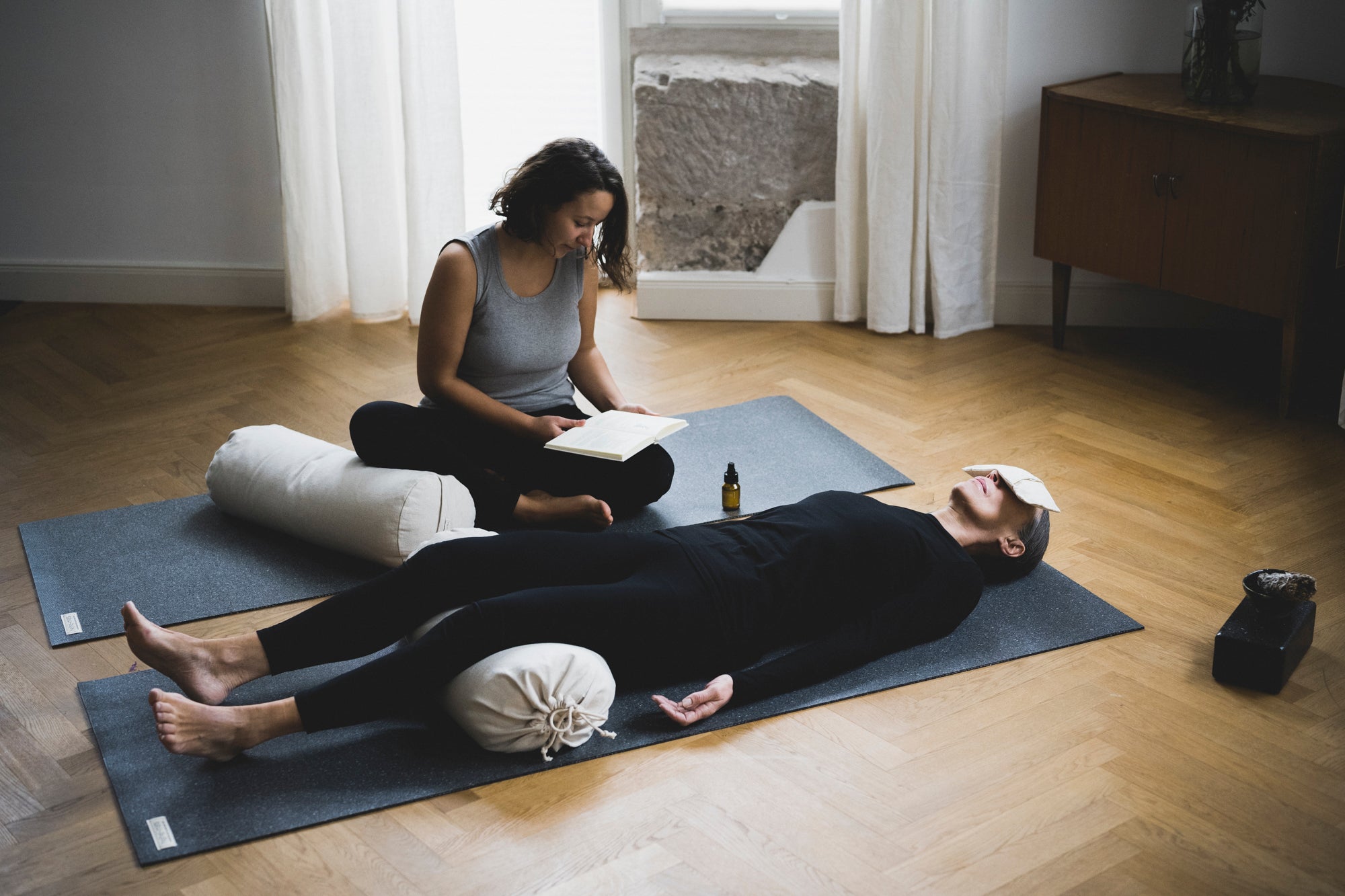 Die Yogalehrerin liest aus einem Buch vor damit die Schülerin entspannen kann mit dem Augenkissen und dem Yoga Bolster