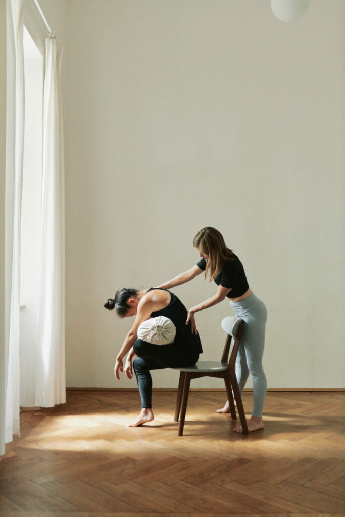 A woman is doing a forward bend on a chair and has the hejhej-bolster under her upper body for support.