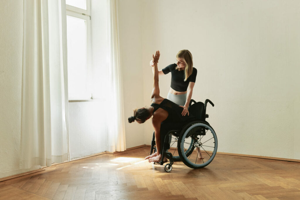 A woman does yoga in her wheelchair and is supported by another woman.